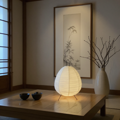 Lighted woven lamp on a wooden table with a framed artwork and vase in the background